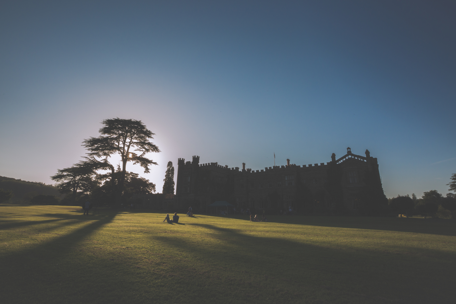 The sun sets over a castle, people sitting on the lawn in the forefround.