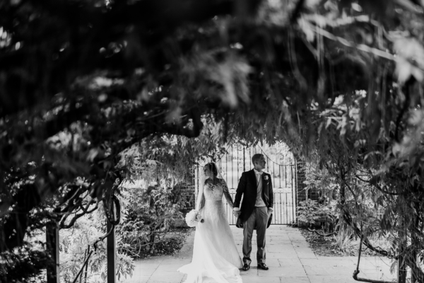 Black & white image of couple standing under wisteria arch