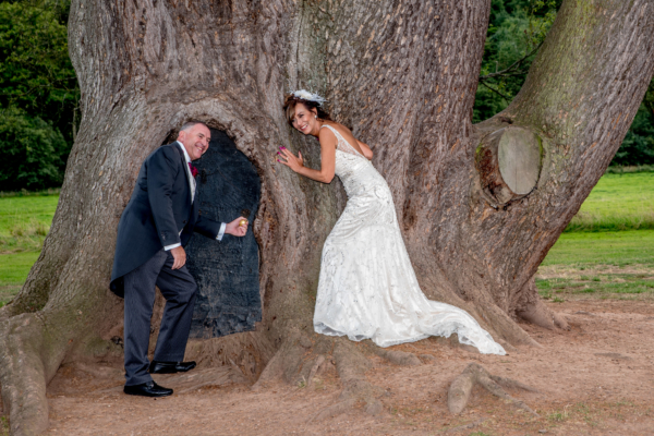 Couple stand by large tree pretending to open small door and listening to the tree.