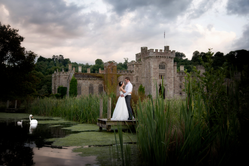 A bride and groom embrace in front of a castle. A swan and cygnet are watching from the lake.