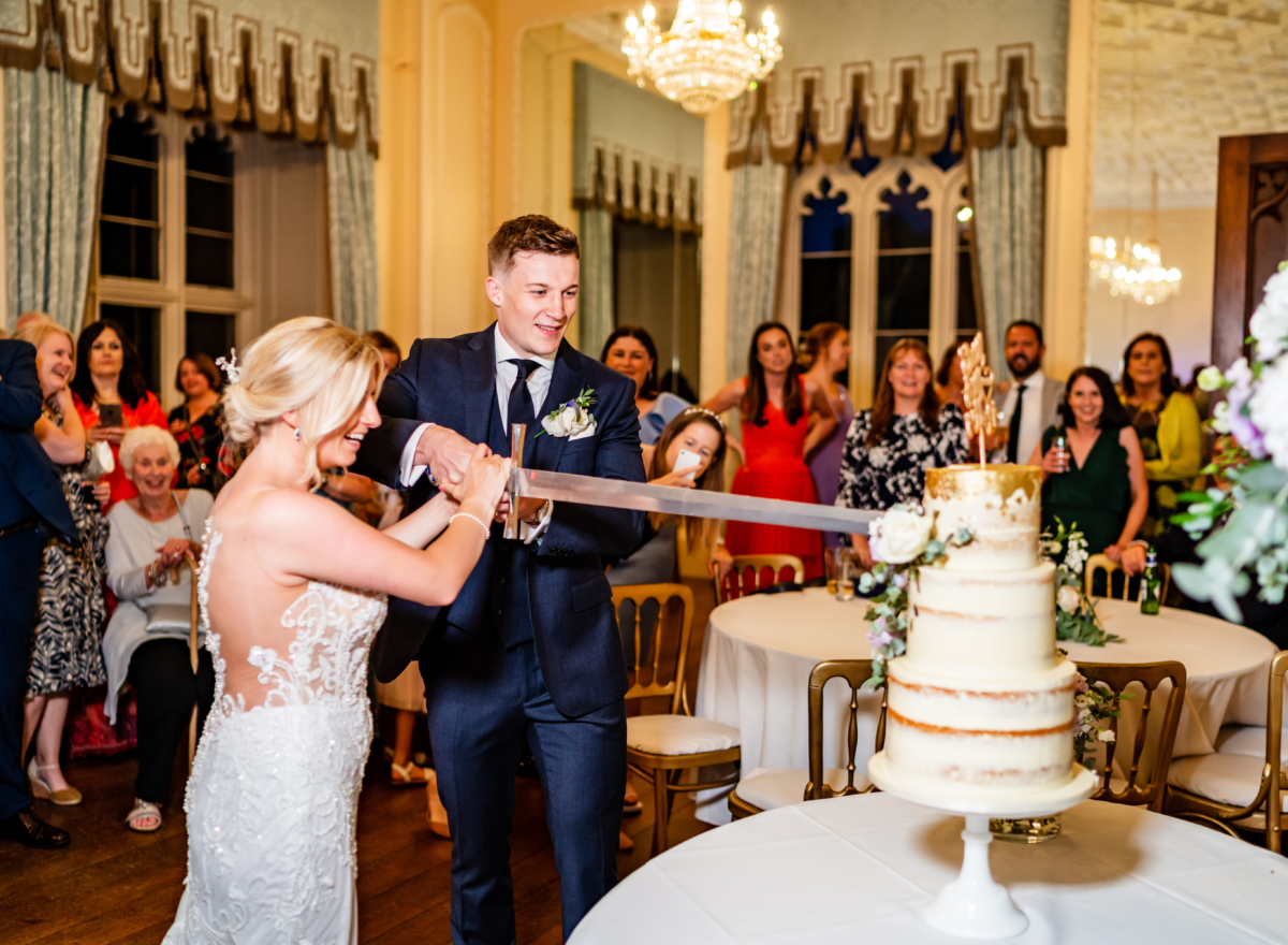 Couple cutting wedding cake in ballroom with sword