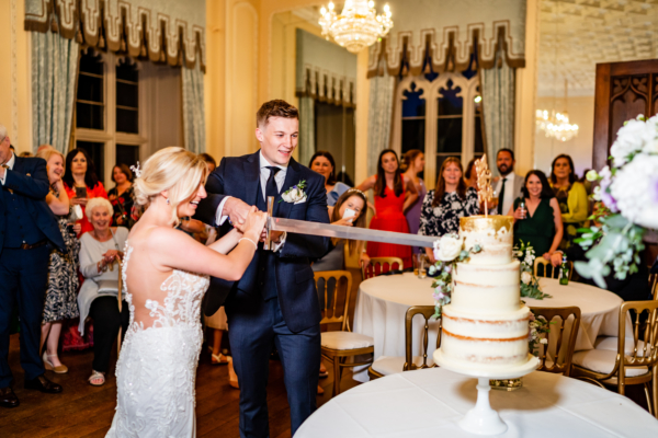 Couple cutting wedding cake in ballroom with sword